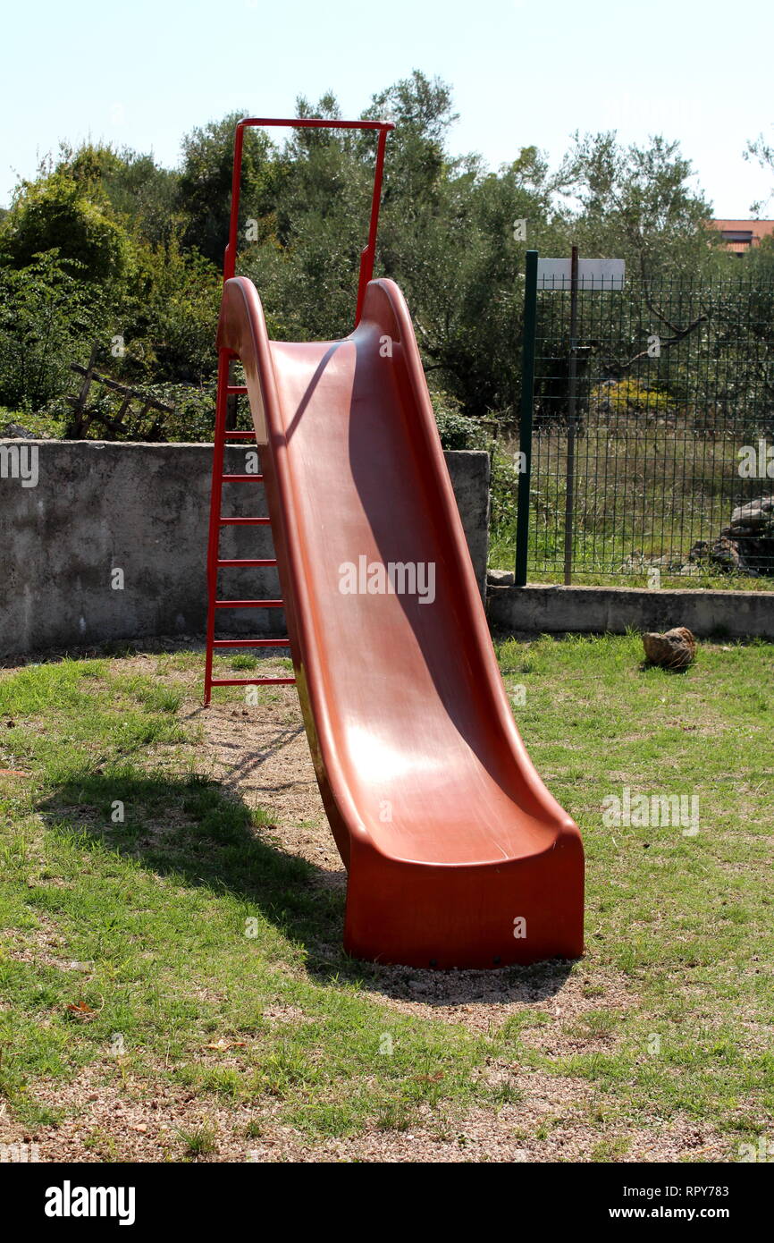 Vintage retrò rosso vecchio parco giochi in plastica scorrono con scale di metallo circondato da erba e muro di pietra con recinzione metallica in background sul giorno caldo e soleggiato Foto Stock