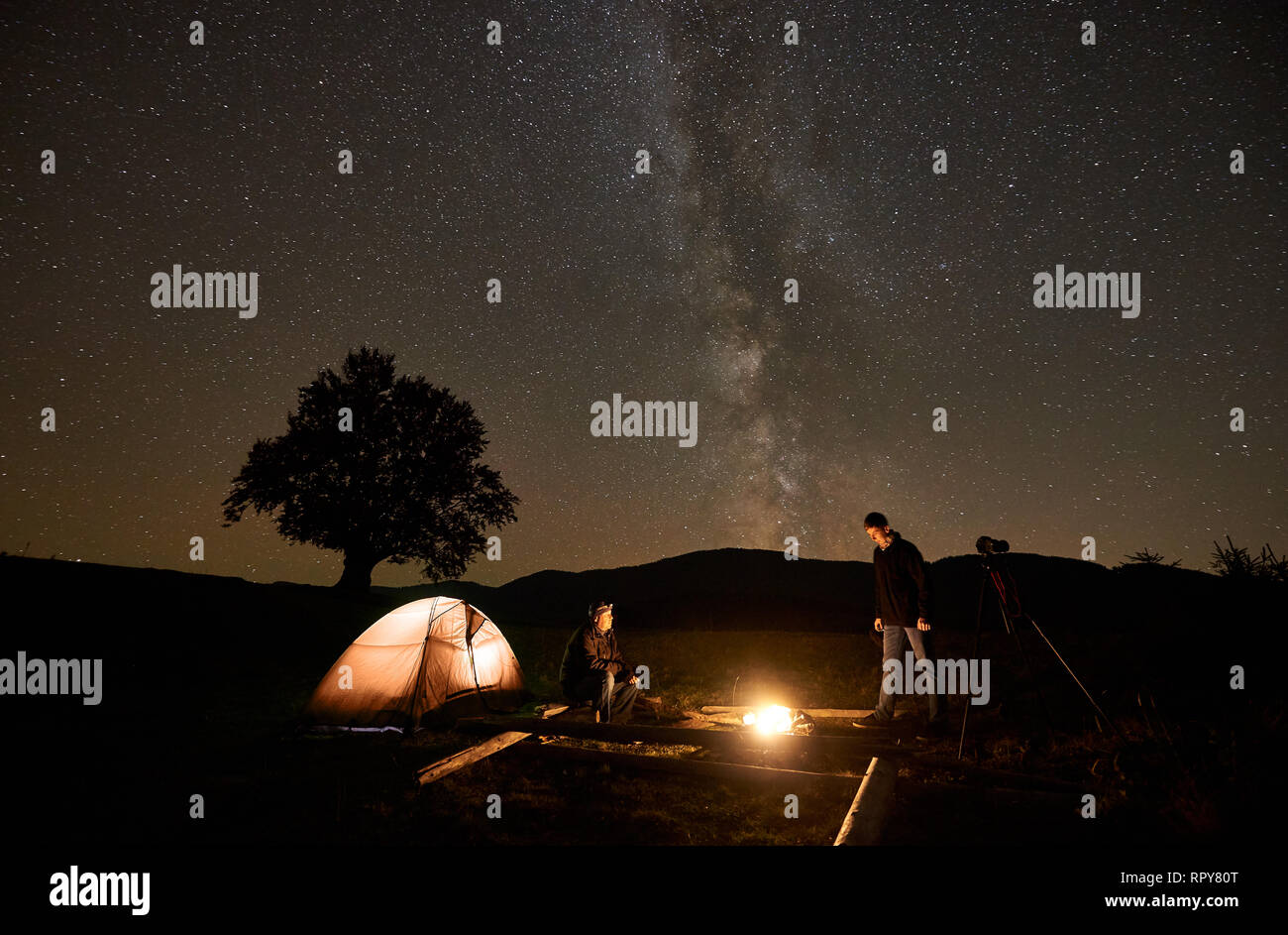 Notte estiva camping, il turismo e la fotografia. Due viaggiatori illuminati da bruciare il falò presso la Tourist tenda e treppiede fotocamera sotto il cielo scuro con scintille luminose Foto Stock