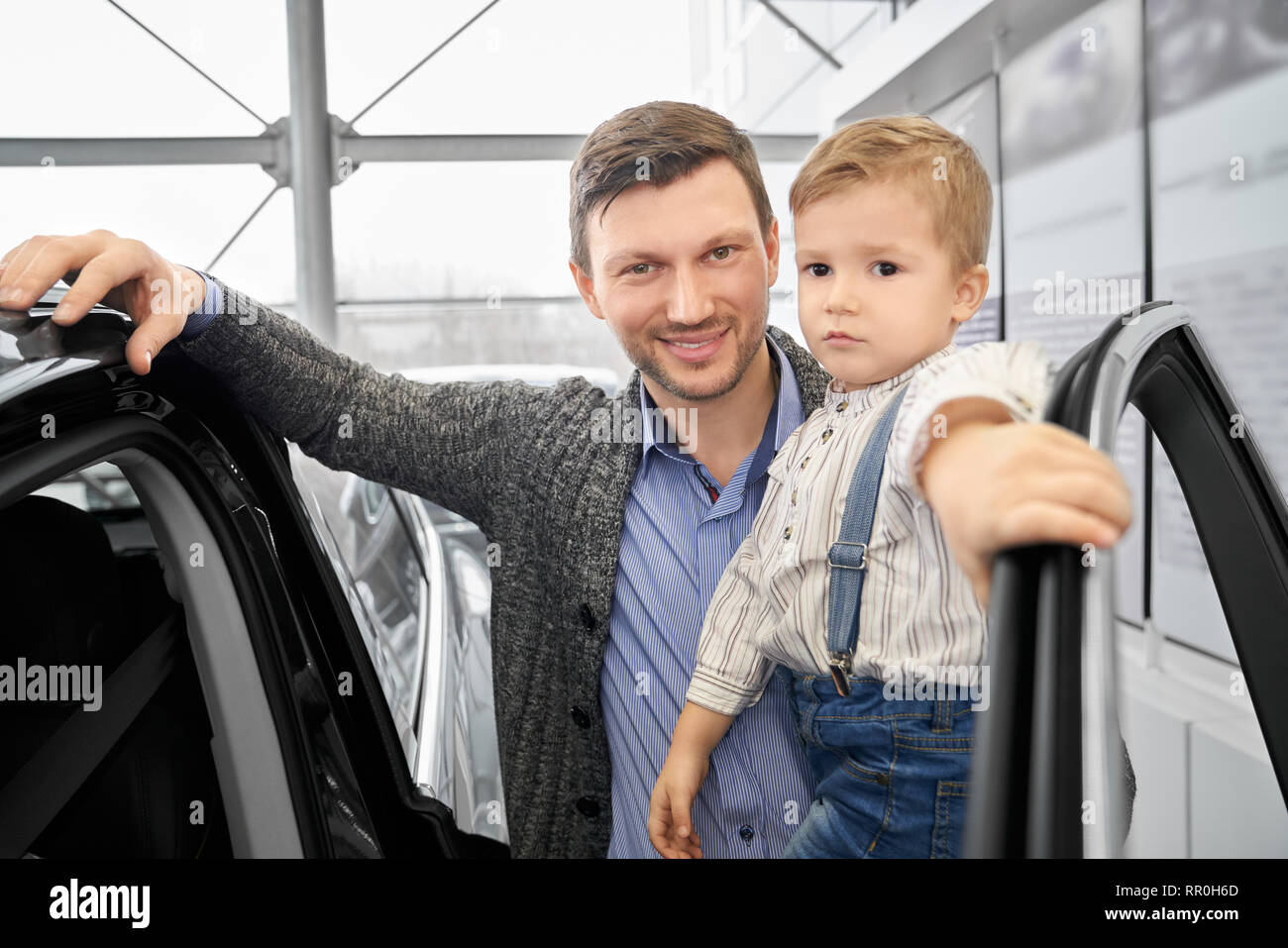 Uomo bello tenendo la mano sulla porta aperta del costoso automobile nero. Allegro padre holding kid a portata di mano. Genitore e bambino sorridente, guardando la telecamera, ponendo in concessionaria auto. Foto Stock