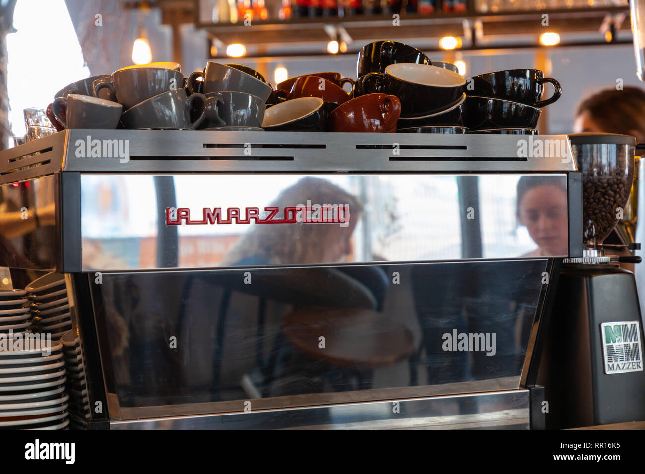 Birmingham, Regno Unito, 24 Febbraio 2019: due donne i capi vengono riflesse in un caffè di argento macchina su un contatore. Le tazze sono impilati sulla parte superiore della macchina. Foto Stock