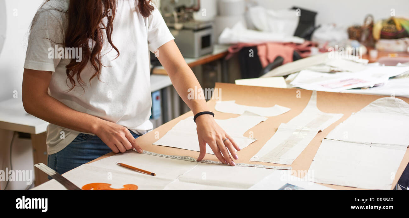 Luogo di lavoro di design studio. Professional designer femmina rendendo i modelli di carta utilizzando nastro di misurazione, righello e la curva su sfondo sfocato di adattare atelier. Vestiti esclusivi Realizzazione concetto. Foto Stock