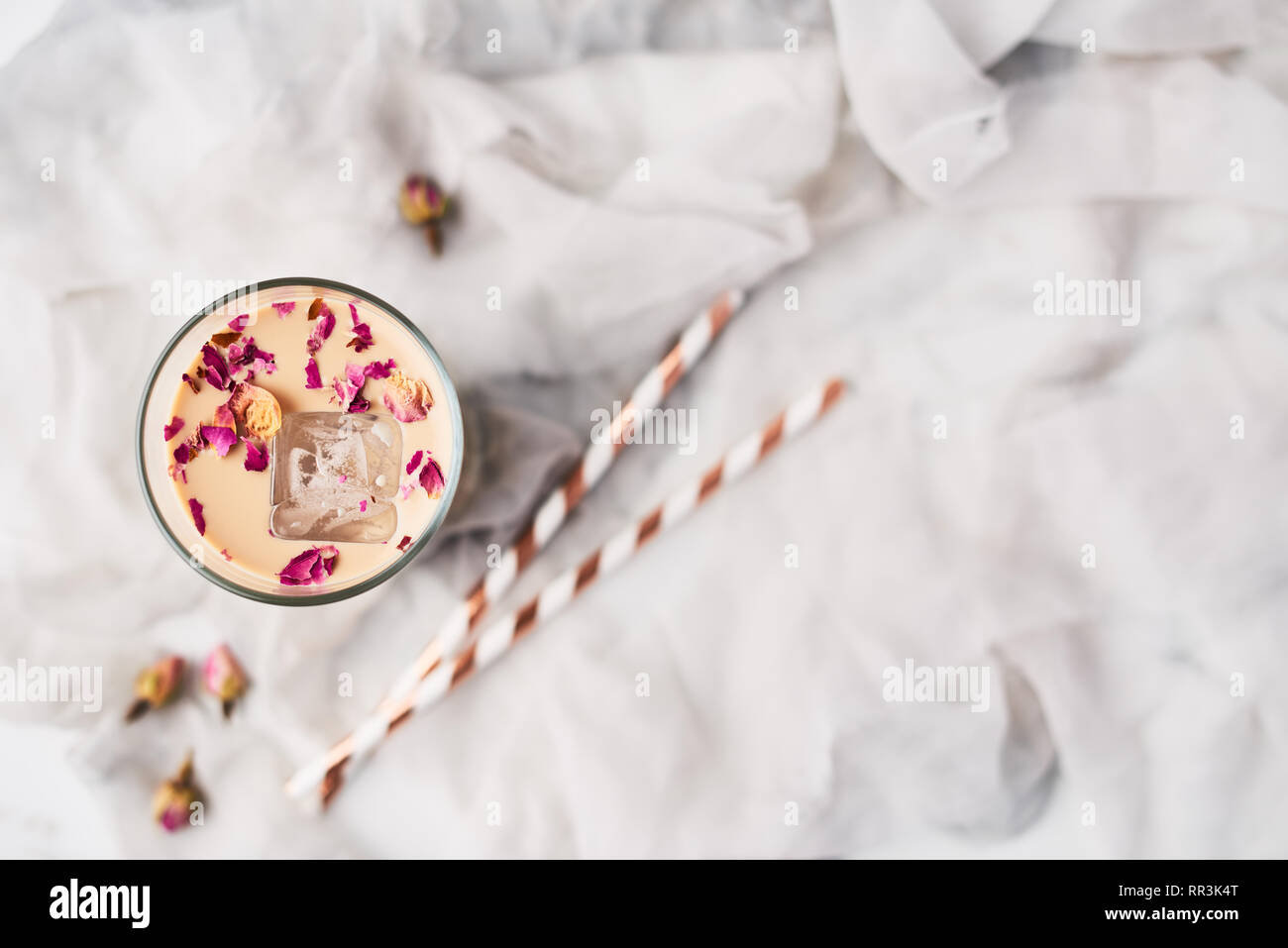 Caffè ghiacciato con rose e cardamomo in un bicchiere alto circondato da rosa appassita e cannucce su bianco e grigio Sfondo di seta. Vista da sopra con spazio copia fo Foto Stock