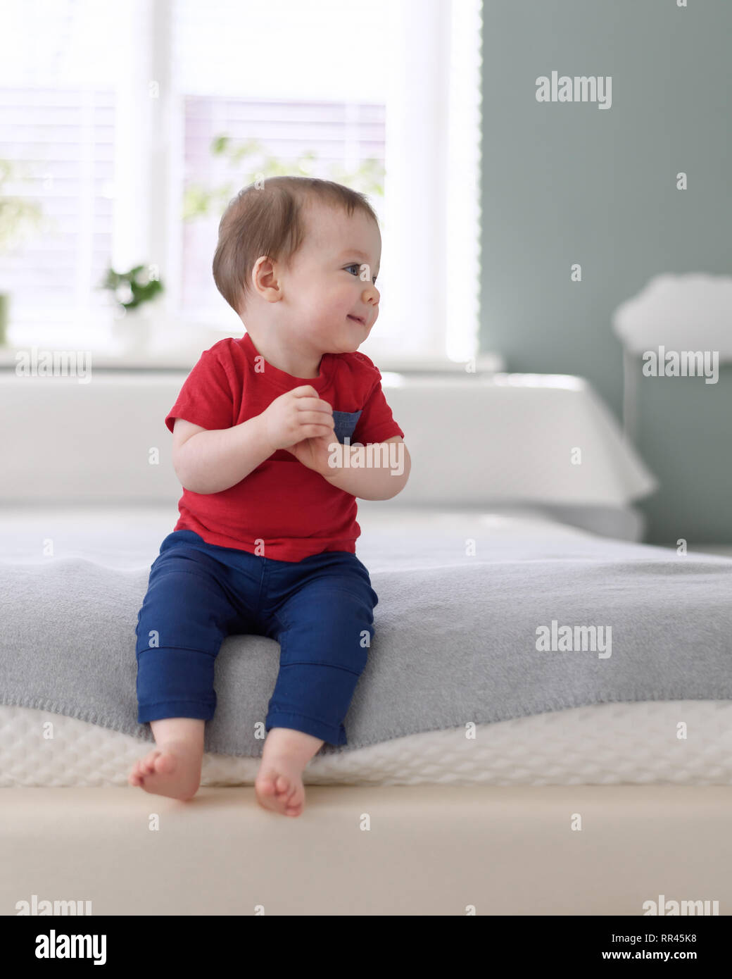 Happy baby boy in maglietta rossa sul letto nella sua camera Foto Stock