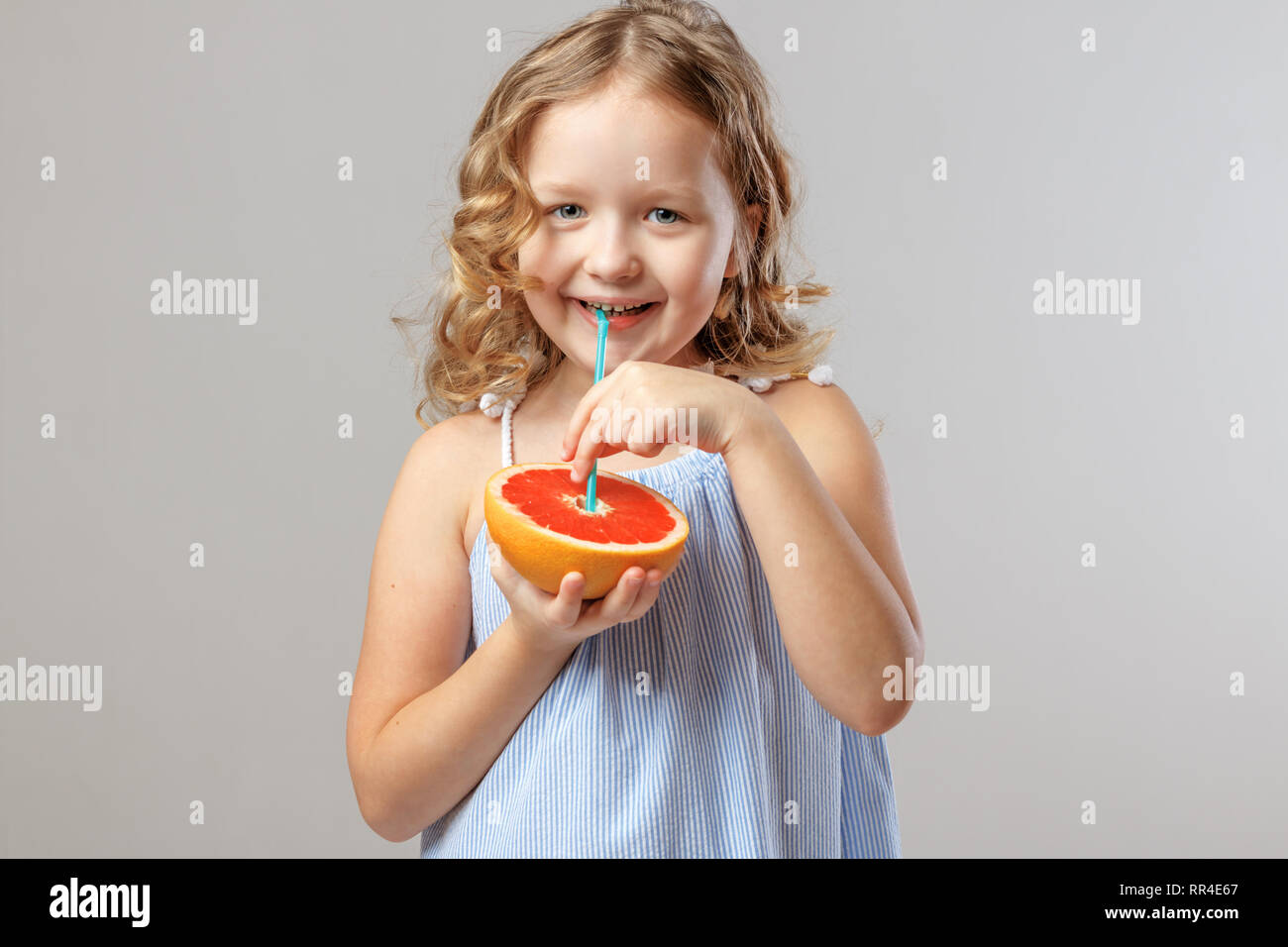 Felice bambina bambino tiene la metà di un pompelmo e beve succo di frutta da un tubulo. Cibo sano. Sfondo grigio, studio Foto Stock
