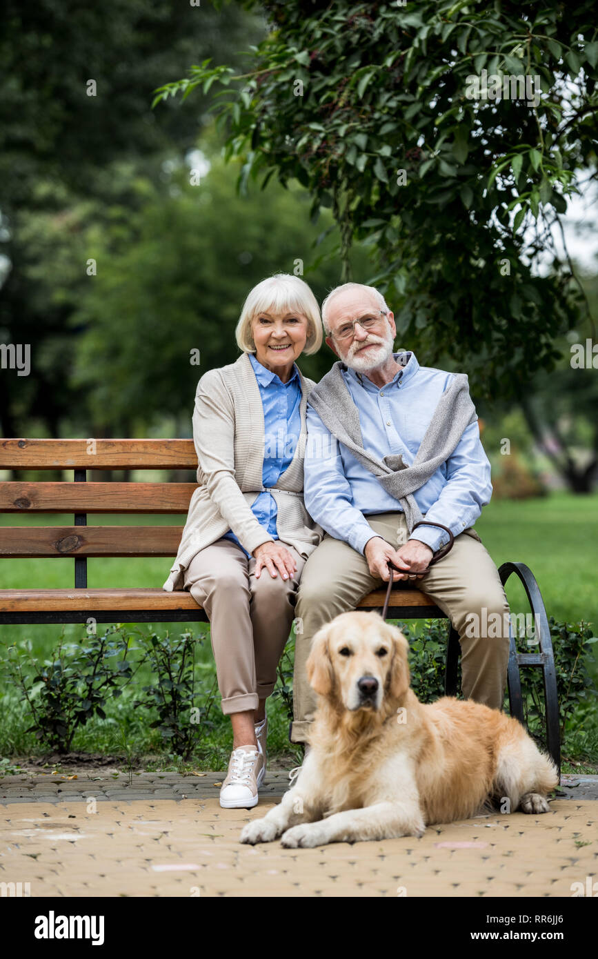 Felice coppia senior seduto sulla panca di legno e cute cane giacenti nelle vicinanze sul marciapiede pavimentato Foto Stock