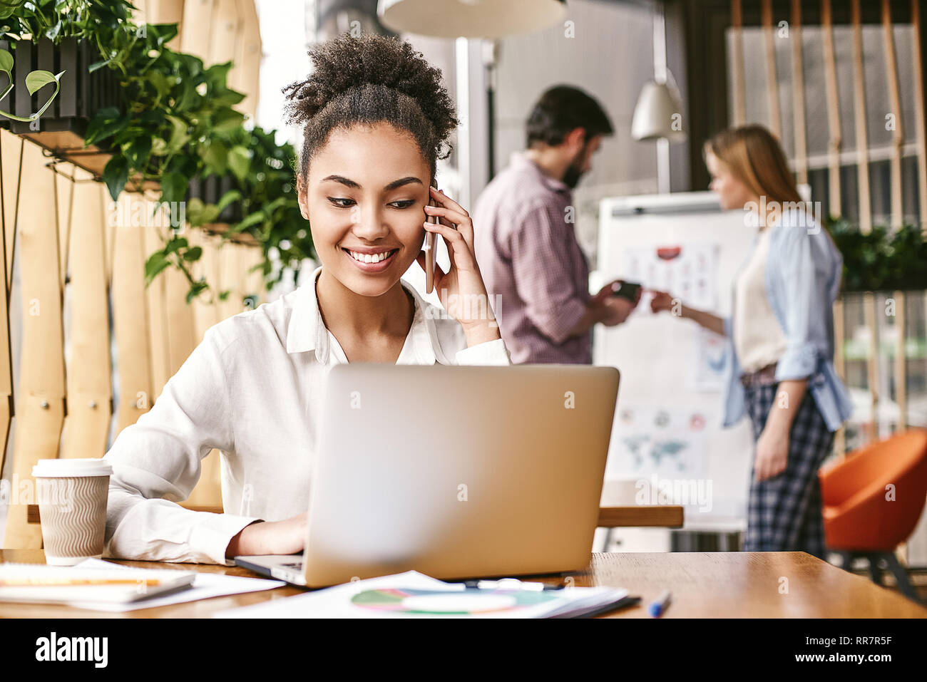 Felice americano africano donna siede in ufficio in legno scrivania, lavorando al computer portatile, avente piacevole chiamata cella o di conversazione. Affascinante ragazza sorride mentre parla sullo smartphone al posto di lavoro. Foto Stock