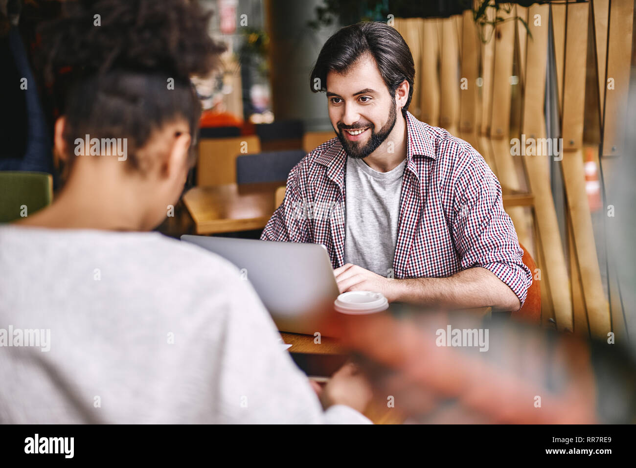 Due allegro colleghi in ufficio indossando abiti casual co-lavorando mentre si utilizza il computer portatile. Barbuto dai capelli scuri cerca uomo al suo collega di sesso femminile con un sorriso mentre lei si concentra sul suo lavoro. Foto Stock