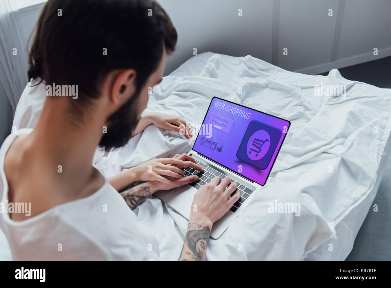 Vista posteriore del giovane giacente in letto, tenendo le mani e utilizzando computer portatile con shopping on line sito web sullo schermo Foto Stock