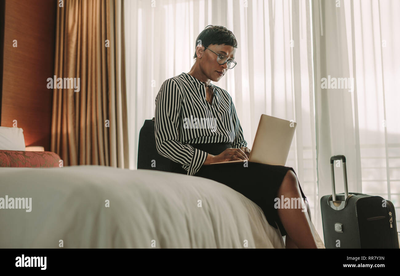 Imprenditrice seduta sul letto tramite computer portatile. Donna africana in viaggio d'affari lavorare in camera d'albergo. Foto Stock
