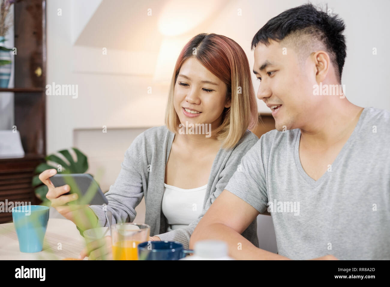 Felice coppia asiatica in pigiama seduti al tavolo in cucina e guardare video sul cellulare togerther a casa la mattina e avente la prima colazione di cereali Foto Stock