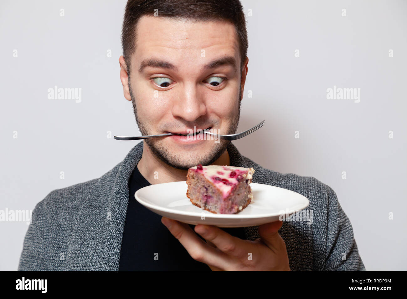 Closeup ritratto giovane carino uomo caucasico sorriso, tenere la forcella nel suo denti e bianco piastra rotonda con un pezzo di torta di biscotti, preparare da mangiare e grin Foto Stock