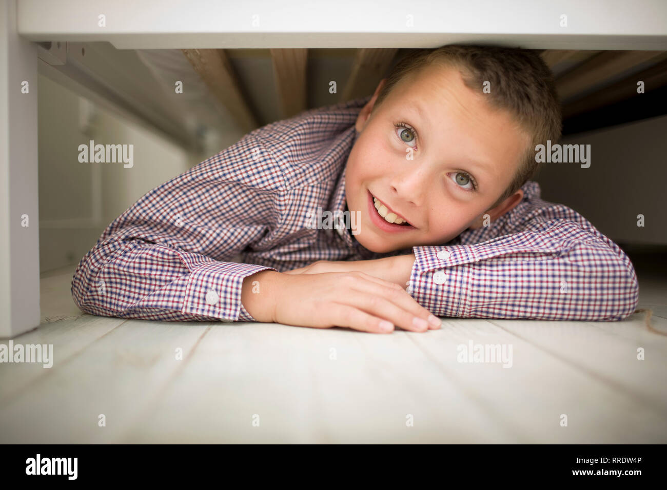 Ritratto di un ragazzo sorridente giacente sul suo stomaco mentre si nasconde sotto un letto in legno. Foto Stock
