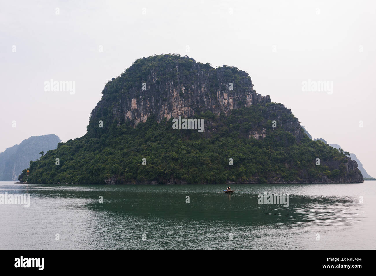 Una piccola barca a remi di fronte a una grande pietra calcarea isola carsica su una mattina nuvoloso, la baia di Ha Long, Vietnam Foto Stock