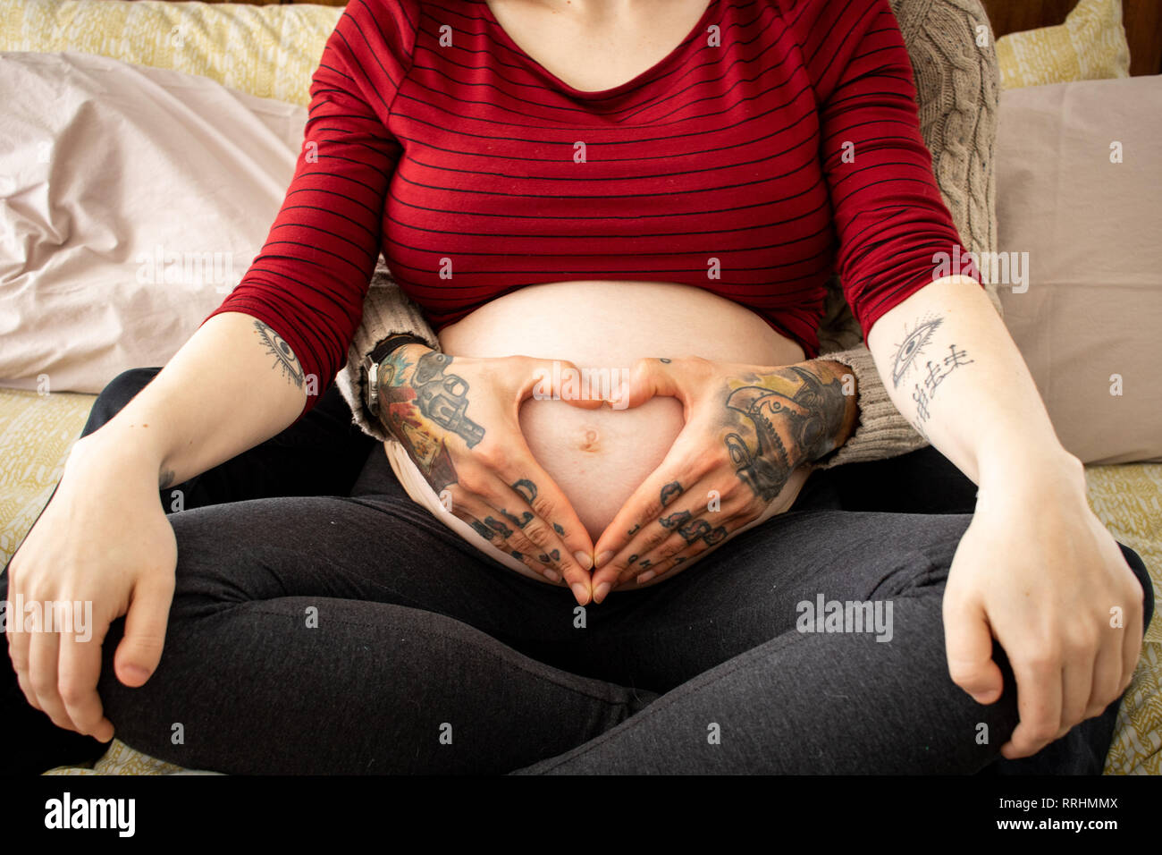 Le sue mani tatuate fanno un cuore sul ventre incinto Foto Stock