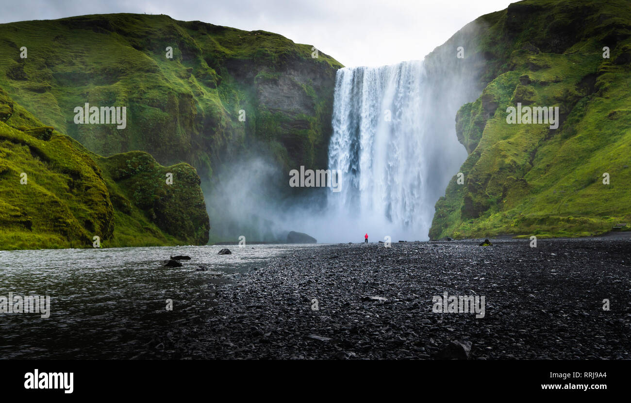 Visitatore sopraffatte da cascate di Skogafoss, Islanda più iconico cascata, situato sul fiume Skoga, Regione meridionale Islanda, regioni polari Foto Stock