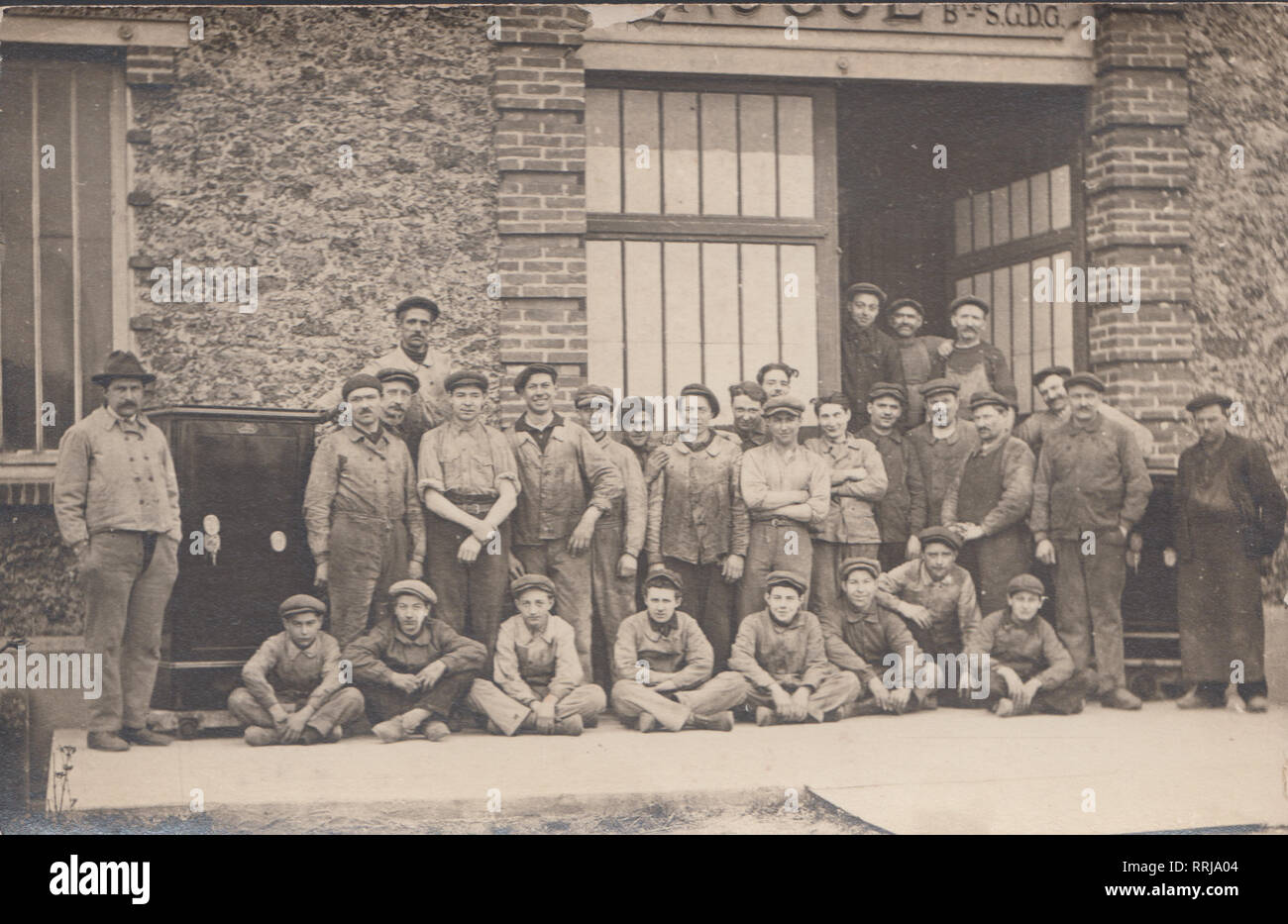 Vintage Cartolina fotografica che mostra un grande gruppo di uomini e ragazzo di lavoratori presso la loro azienda di produzione. Grande Cassaforte visibile. Foto Stock