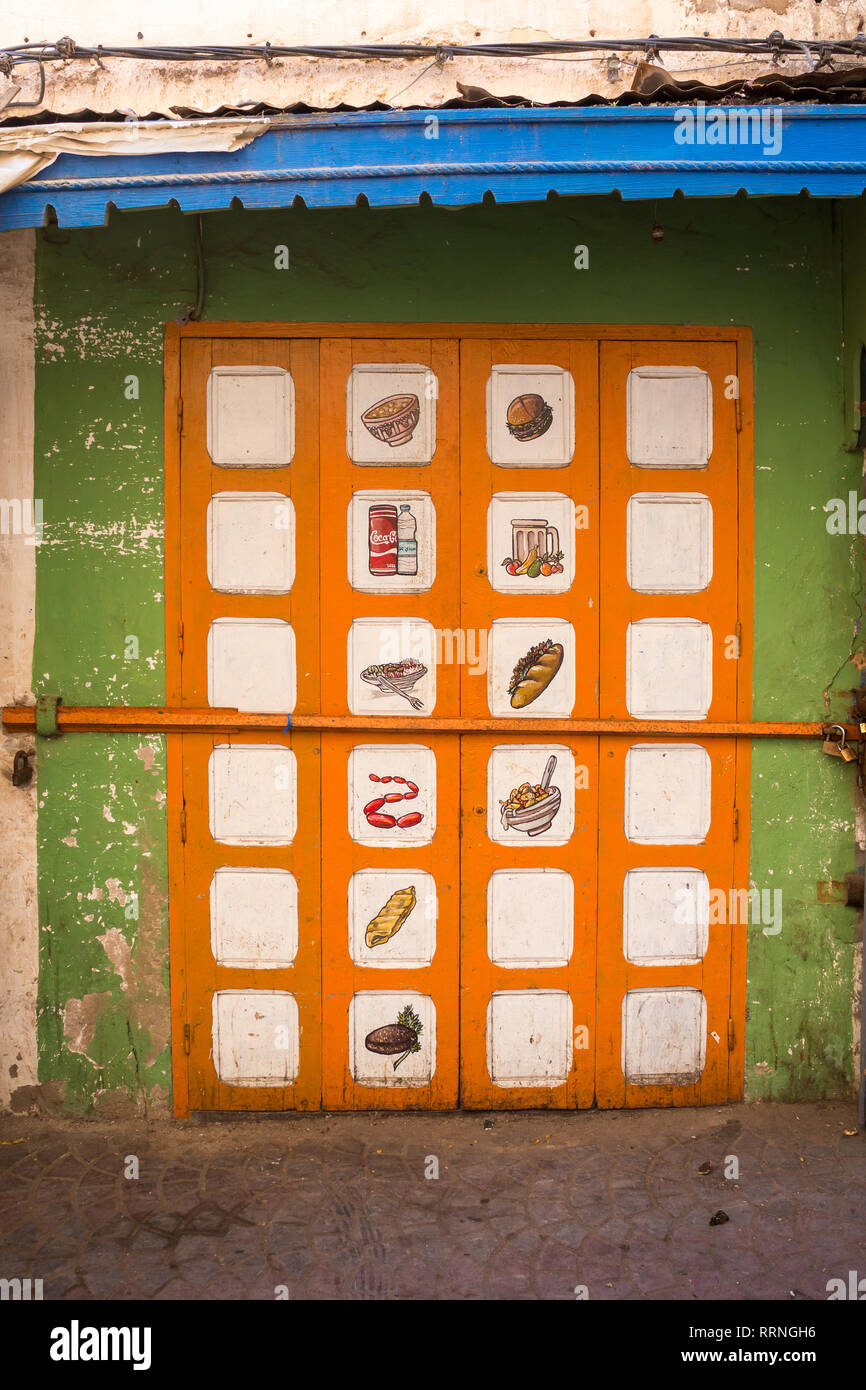 Parete verde e arancione gate di un foodstore chiuso, con le immagini dipinte di vari prodotti alimentari. Marquise blu. Safi, Marocco. Foto Stock