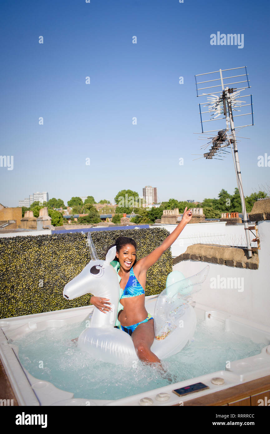 Ritratto giocoso e spensierato giovane donna in bikini su Pegasus gonfiabile nella soleggiata terrazza sul tetto la vasca calda Foto Stock
