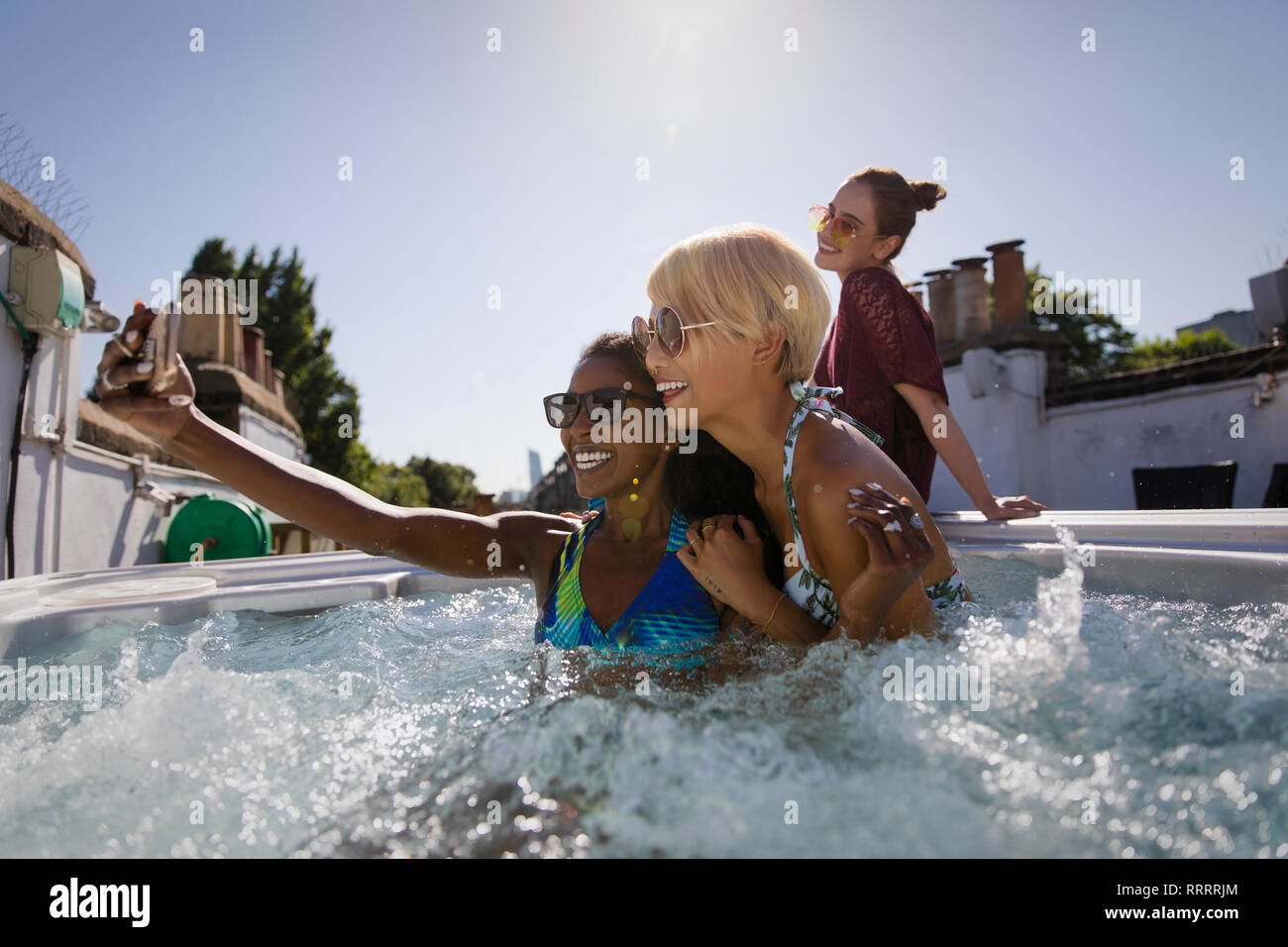 Giovani donne amici prendendo selfie nella soleggiata terrazza sul tetto la vasca calda Foto Stock
