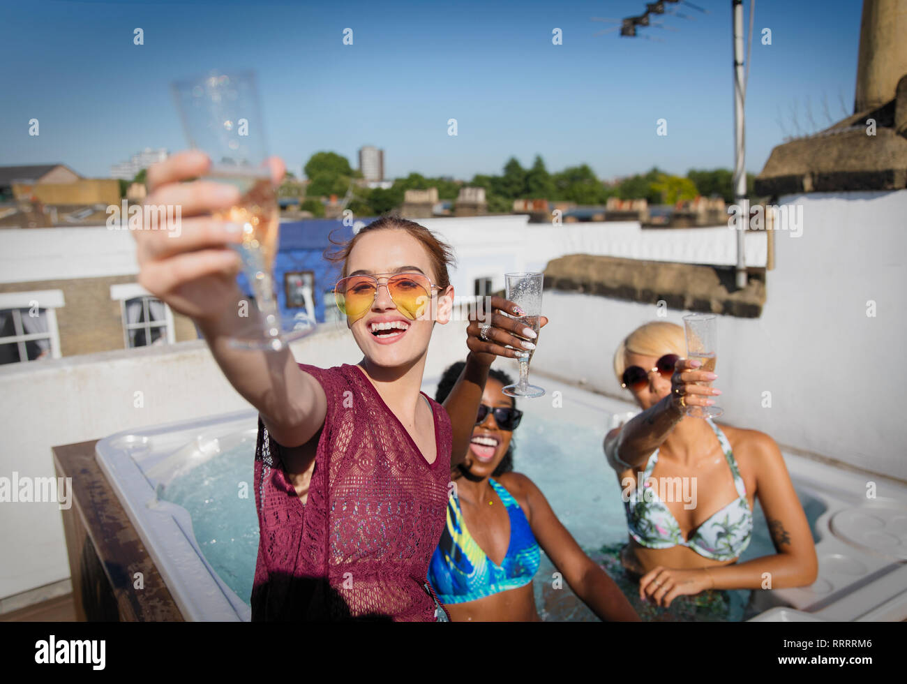 Ritratto fiducioso e spensierato di donne giovani amici bevendo champagne nella soleggiata terrazza sul tetto la vasca calda Foto Stock