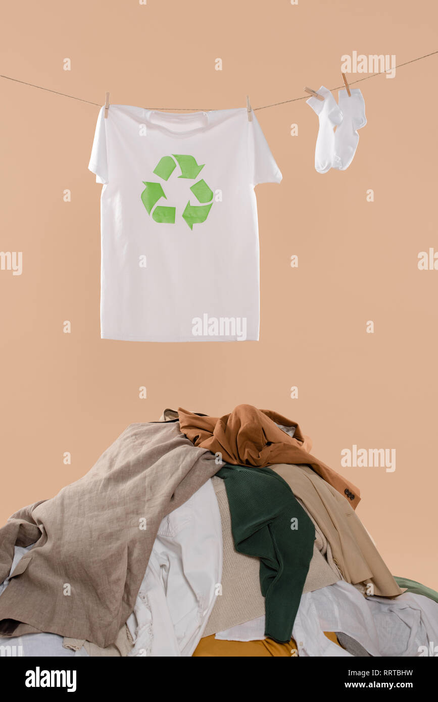 T-shirt con segno di riciclaggio su stendibiancheria vicino calze bianche e pila di capi di abbigliamento su sfondo beige, risparmio ambientale concetto Foto Stock