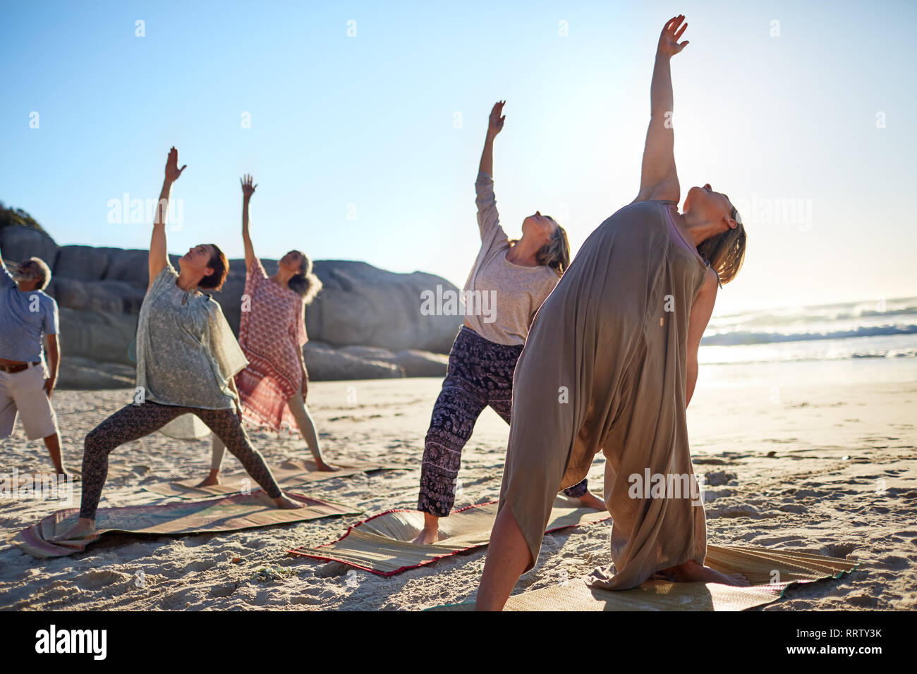 Gruppo a praticare yoga guerriero retromarcia pongono sulla spiaggia soleggiata durante il ritiro di yoga Foto Stock