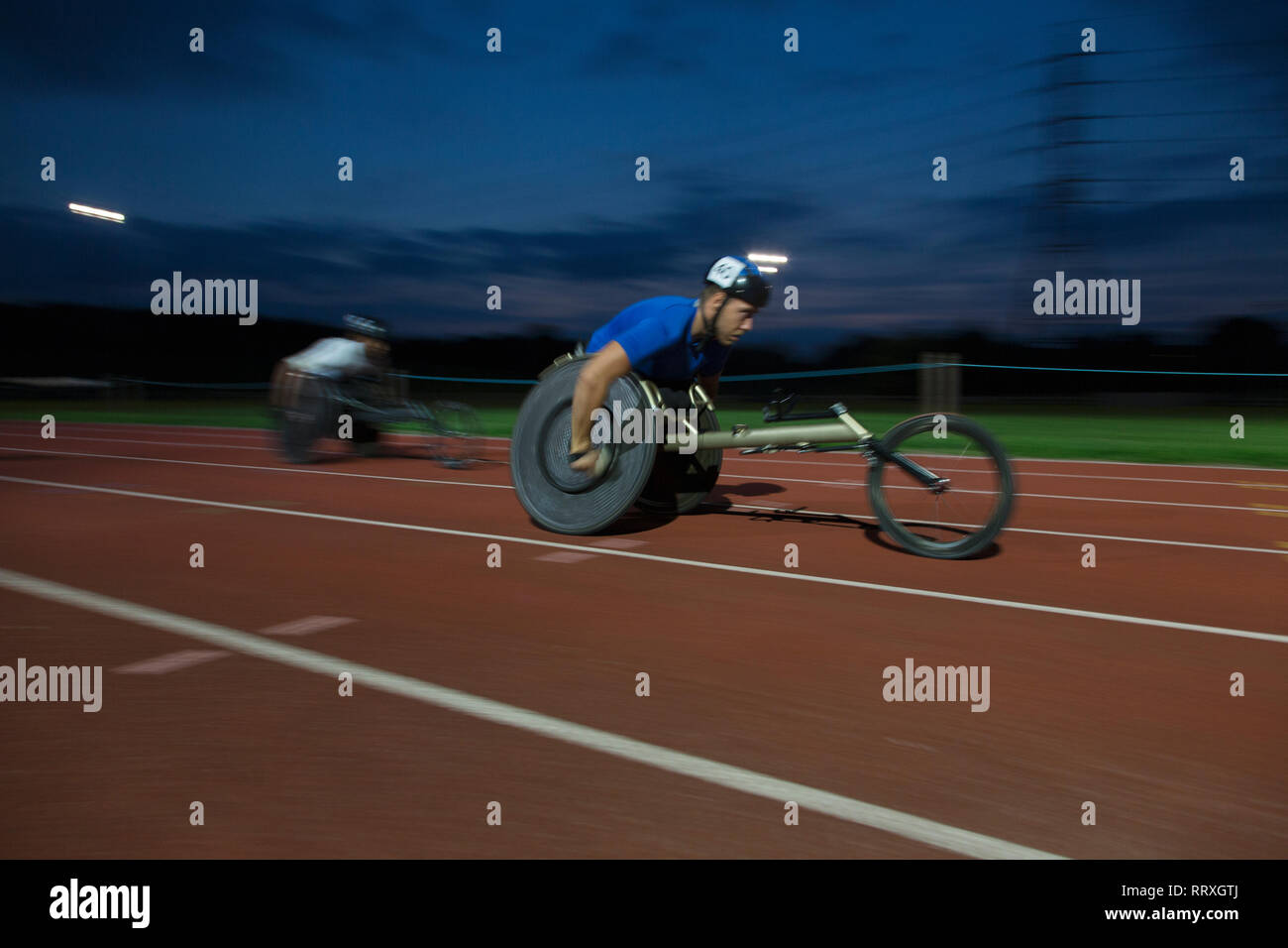 Determinato maschile giovane atleta paraplegico accelerando lungo la via dello sport in carrozzella gara in notturna Foto Stock