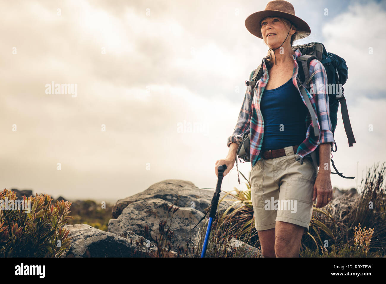 Senior donna che indossa un cappello e uno zaino trekking nella campagna  tenendo un trekking pole. Avventurosa donna su un sentiero escursionistico  in un giorno nuvoloso Foto stock - Alamy