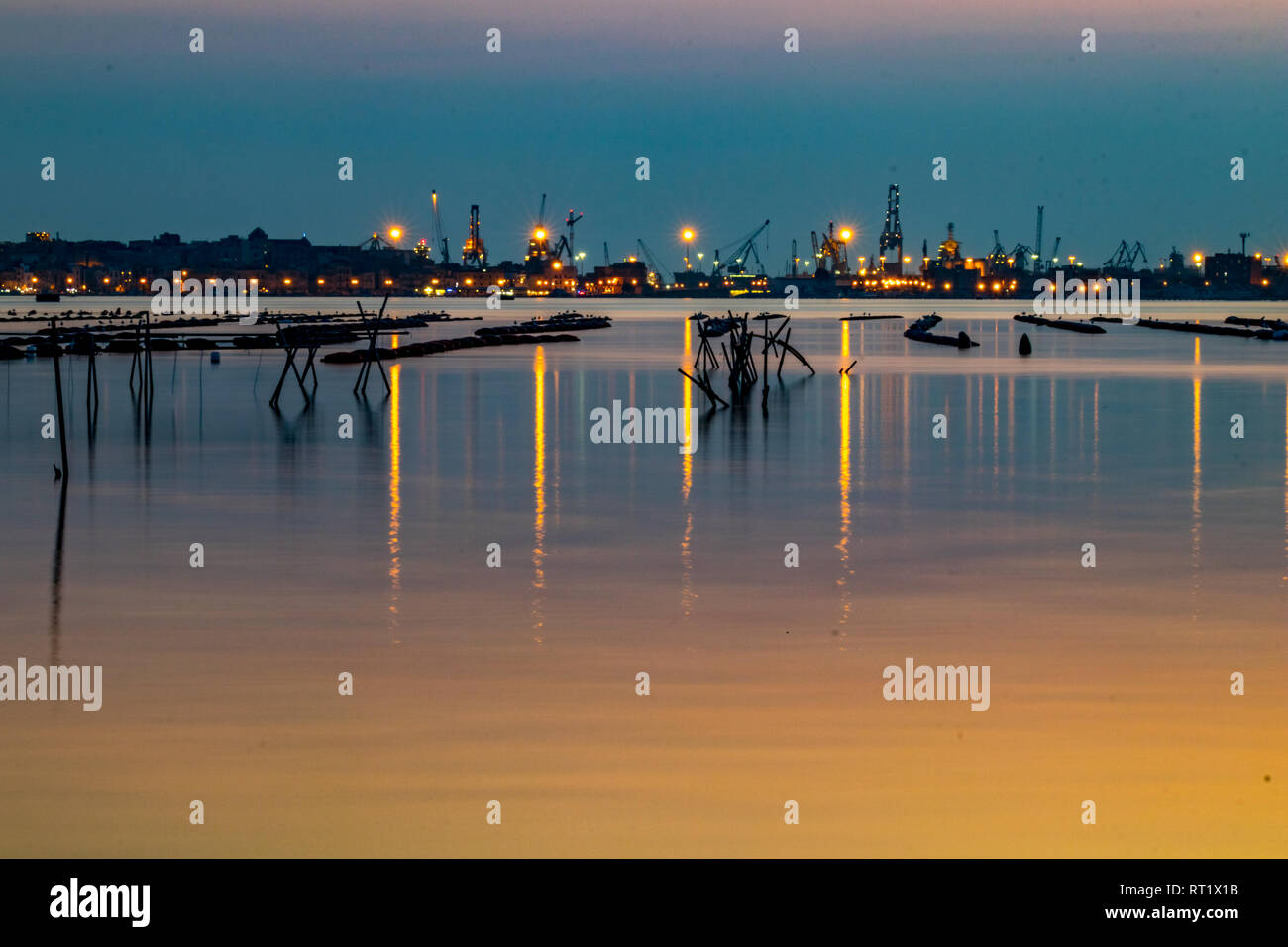 La fotografia notturna, una lunga esposizione immagine del cantiere e la zona industriale di Taranto, Italia con attrezzature di pesca nella parte anteriore e di acqua bella riflessi e colori del tramonto Foto Stock