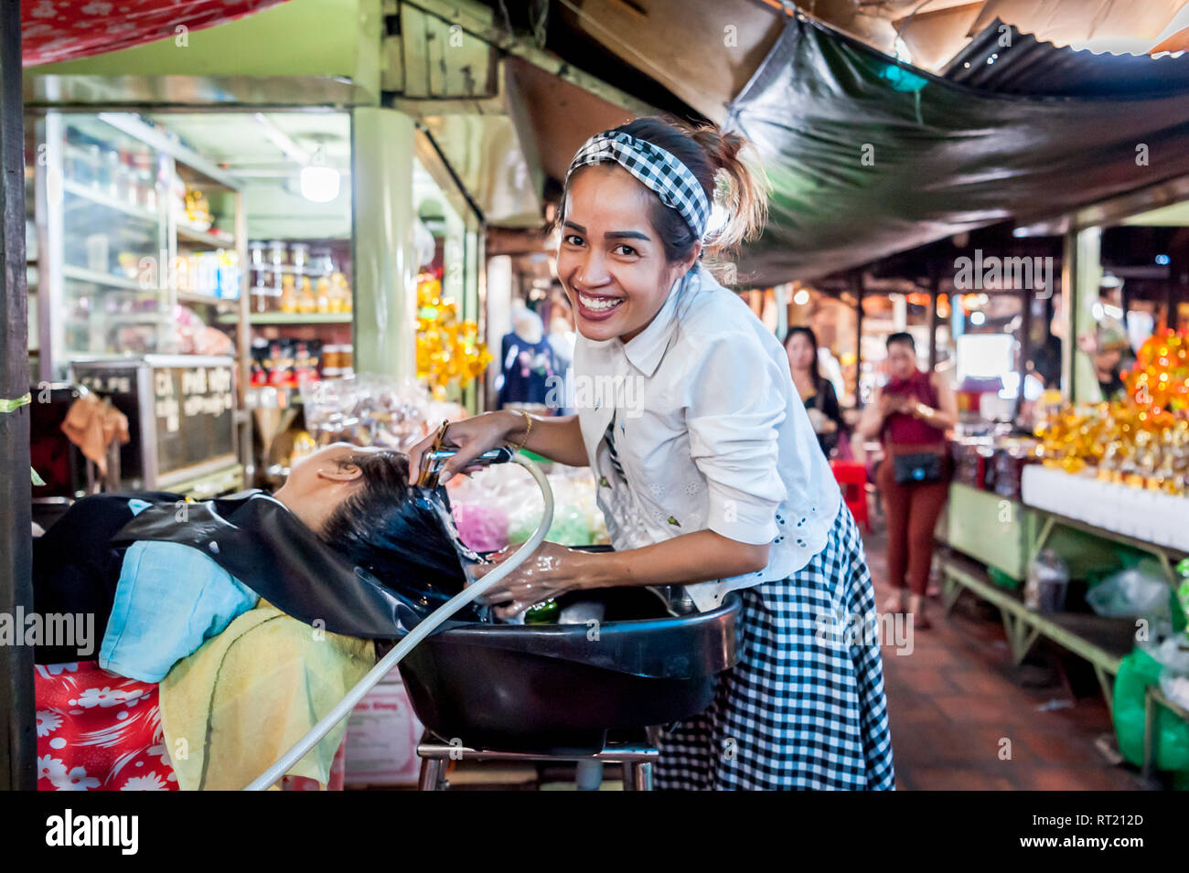 Una signora di lavaggi e tagli di capelli di donne in un parrucchiere in un affollato mercato coperto in Phnom Penh Cambogia. Foto Stock Una signora di lavaggi e tagli di capelli di donne in un parrucchiere in un affollato mercato coperto in Phnom Penh Cambogia. Foto Stock