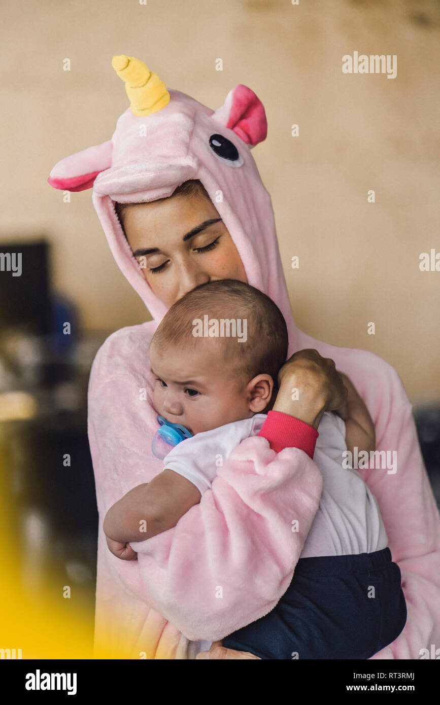 Giovane madre indossa unicorn onesie, stando in ufficio, tenendo il suo figlio nelle sue braccia Foto Stock
