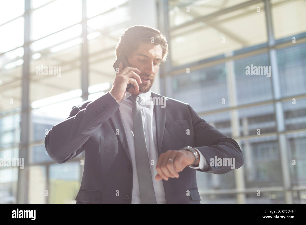 Imprenditore tempo di controllo mentre si parla al telefono cellulare in ufficio lobby Foto Stock