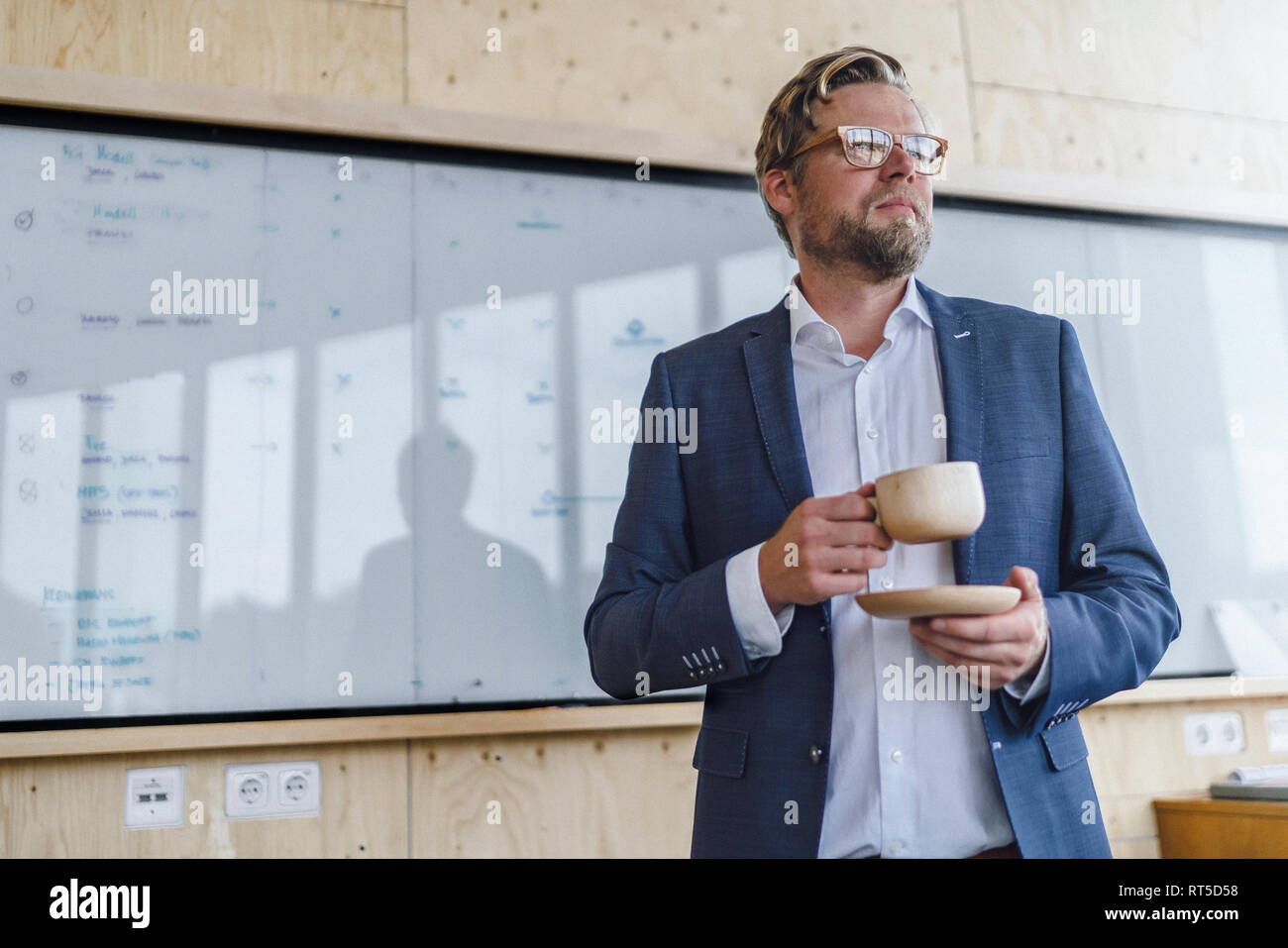 Imprenditore in piedi nel suo ufficio a fronte della presentazione con lavagna, bere il caffè da una tazza di legno Foto Stock