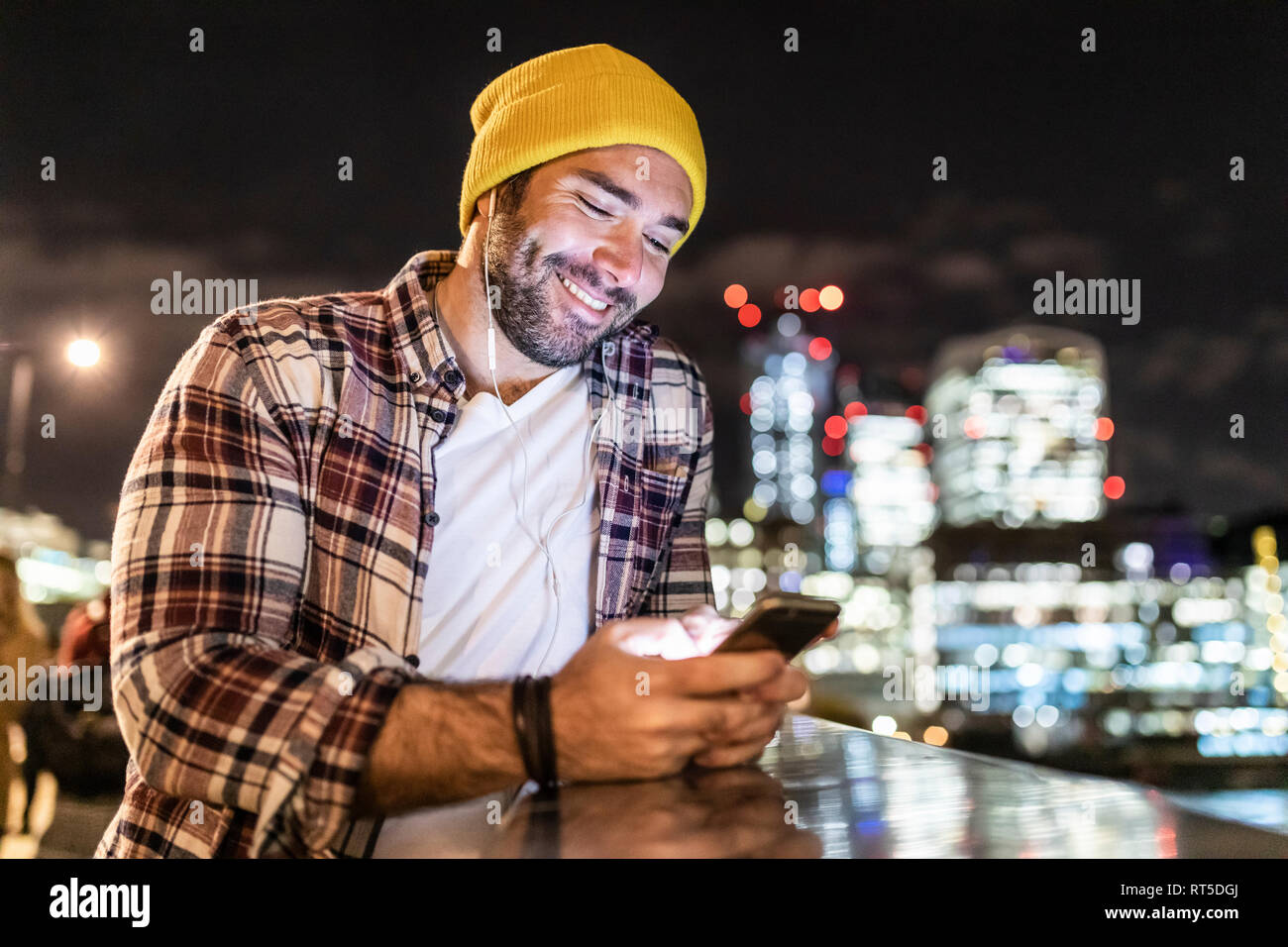 UK, Londra, uomo sorridente appoggiato su di una ringhiera e guardando il suo telefono con le luci della città in background Foto Stock