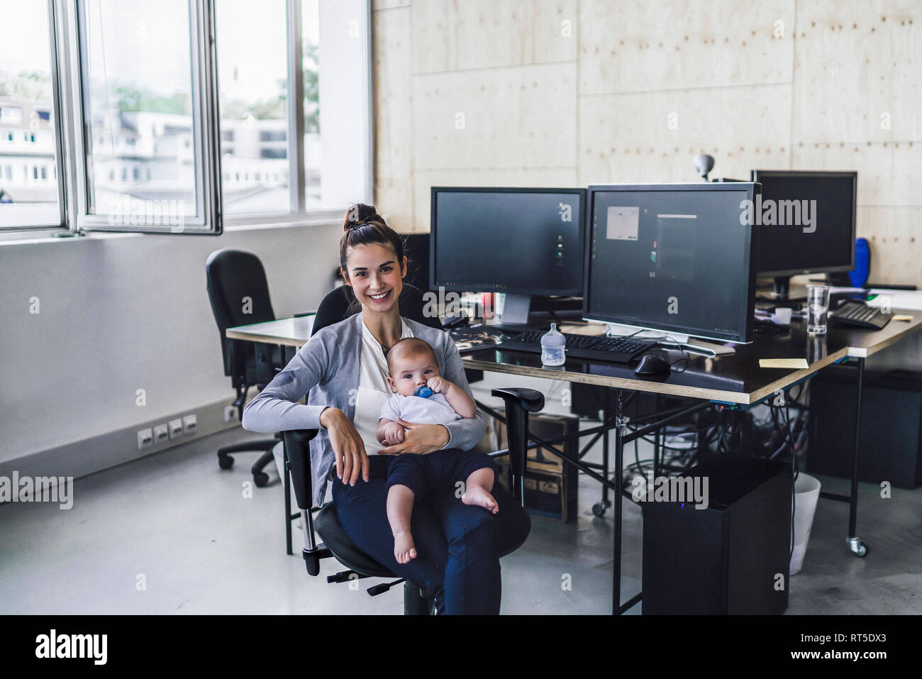 Lavorando madre con bambino sul suo grembo, seduta in ufficio Foto Stock