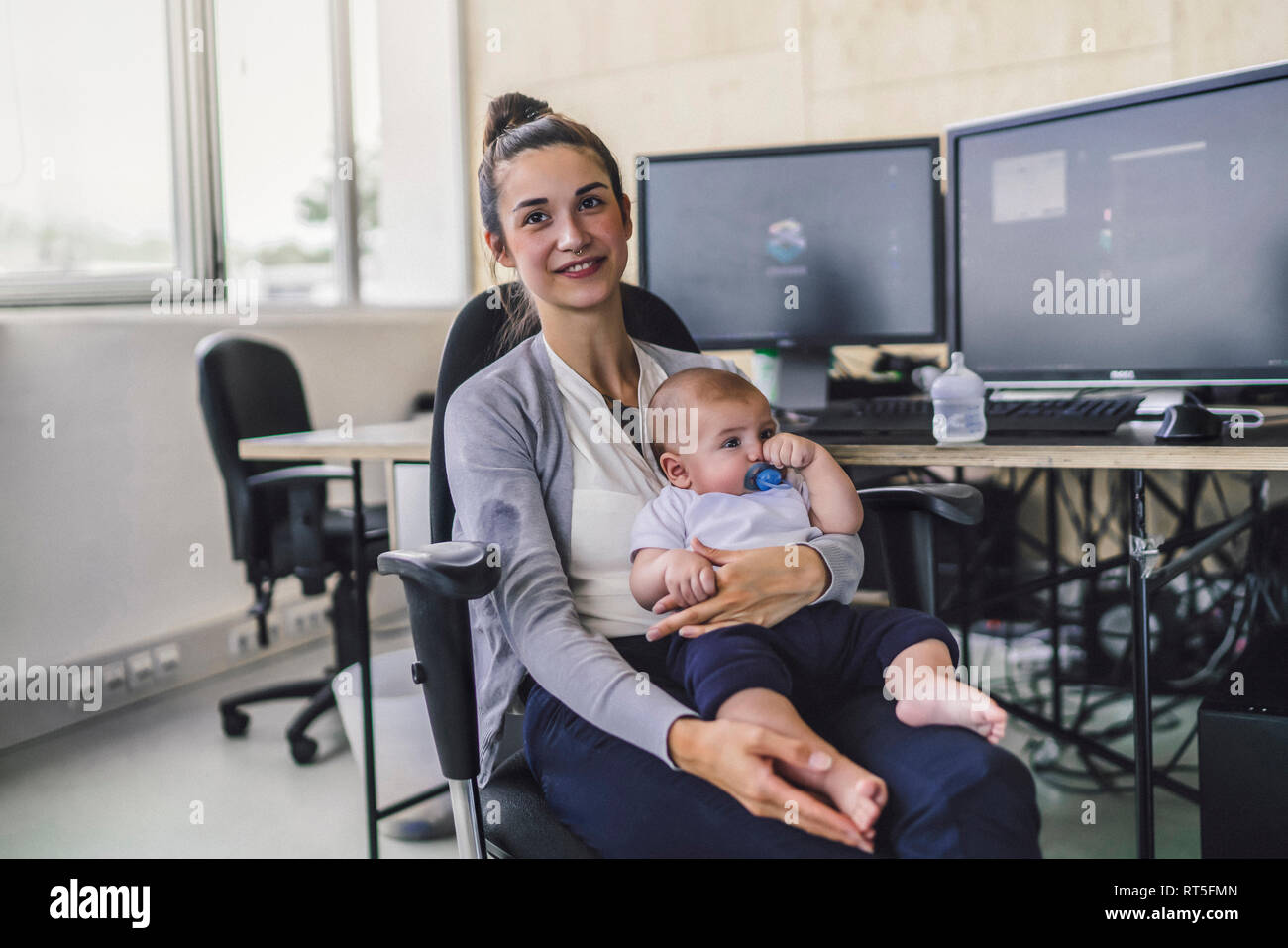 Lavorando madre con bambino sul suo grembo, seduta in ufficio Foto Stock