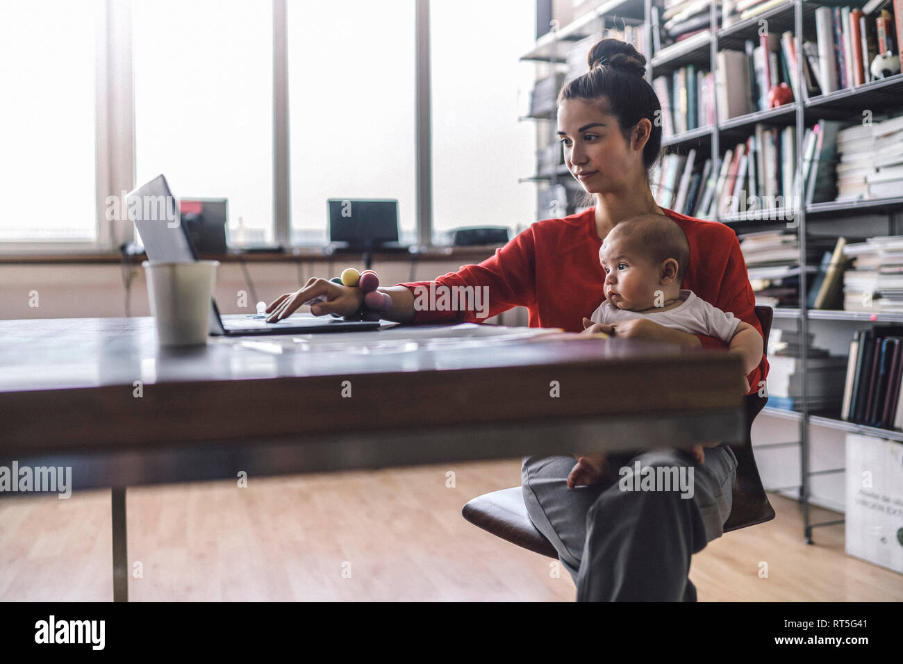 Lavorando madre con bambino sul suo grembo, seduta in ufficio, con il computer portatile Foto Stock