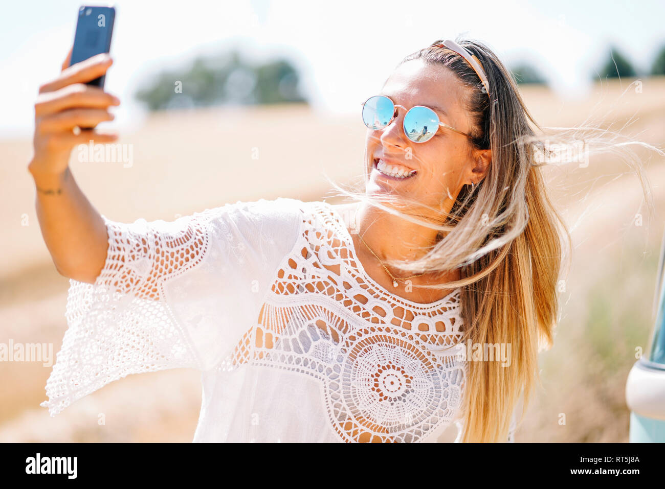 Ritratto di giovane donna bionda indossando occhiali da sole con mirroring tenendo selfie con lo smartphone Foto Stock