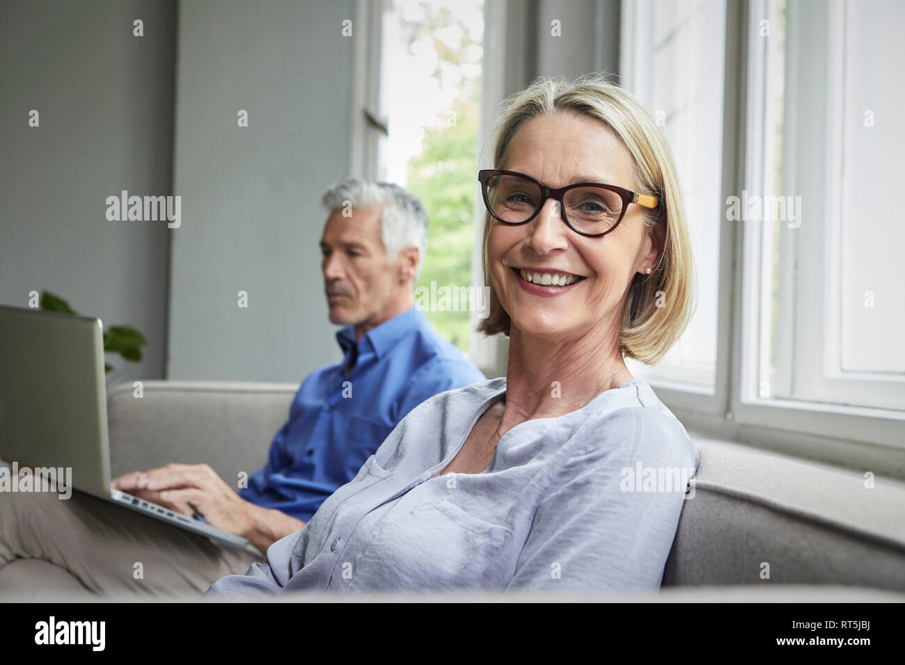 Ritratto di sorridere donna matura sul lettino con uomo in background utilizzando tablet Foto Stock