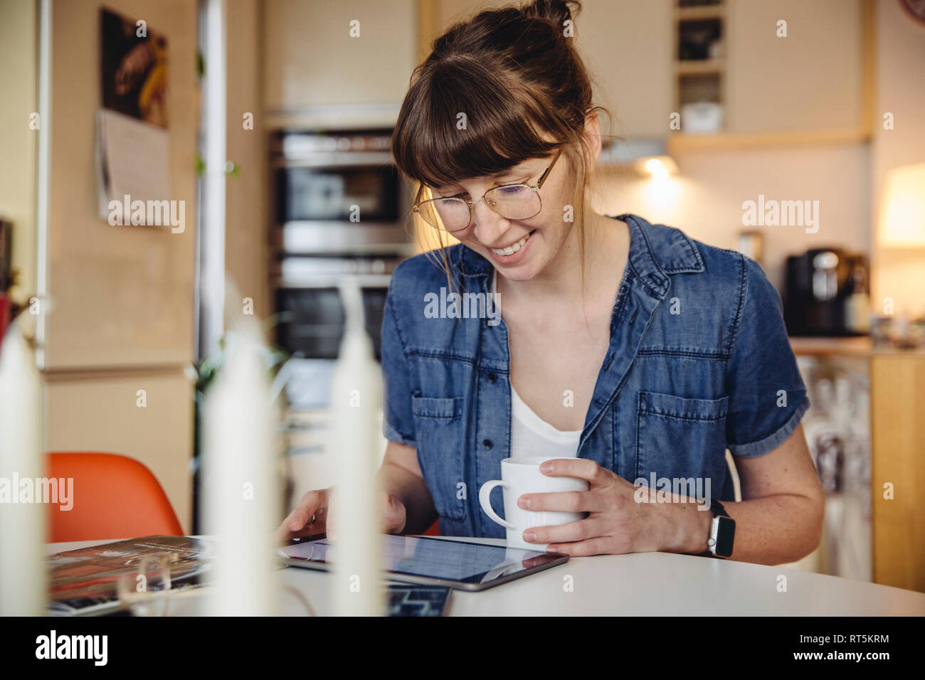 Donna sorridente seduto con tazza di caffè al tavolo in cucina utilizzando tablet Foto Stock