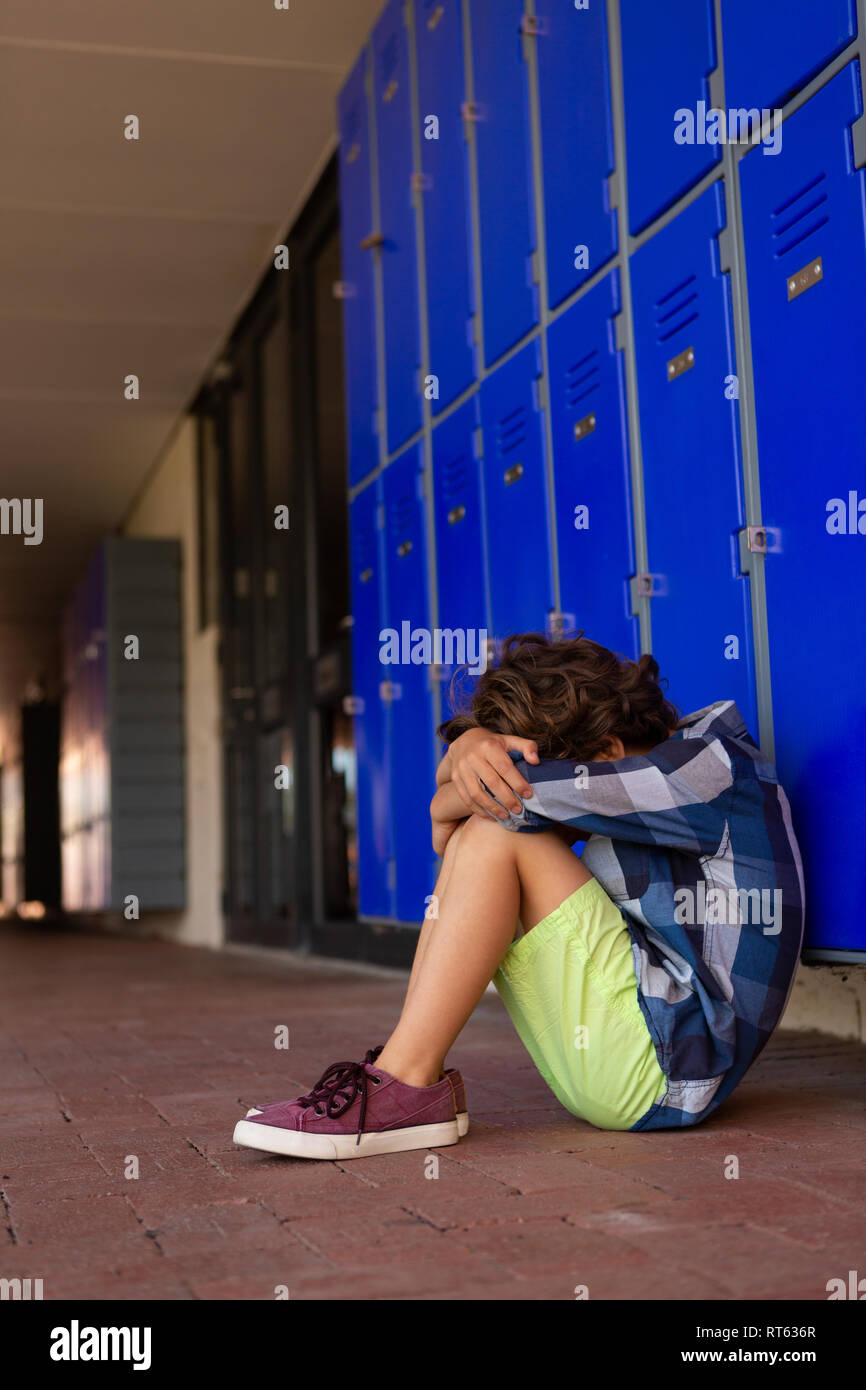 Triste schoolboy seduto da solo sul pavimento nel corridoio Foto Stock