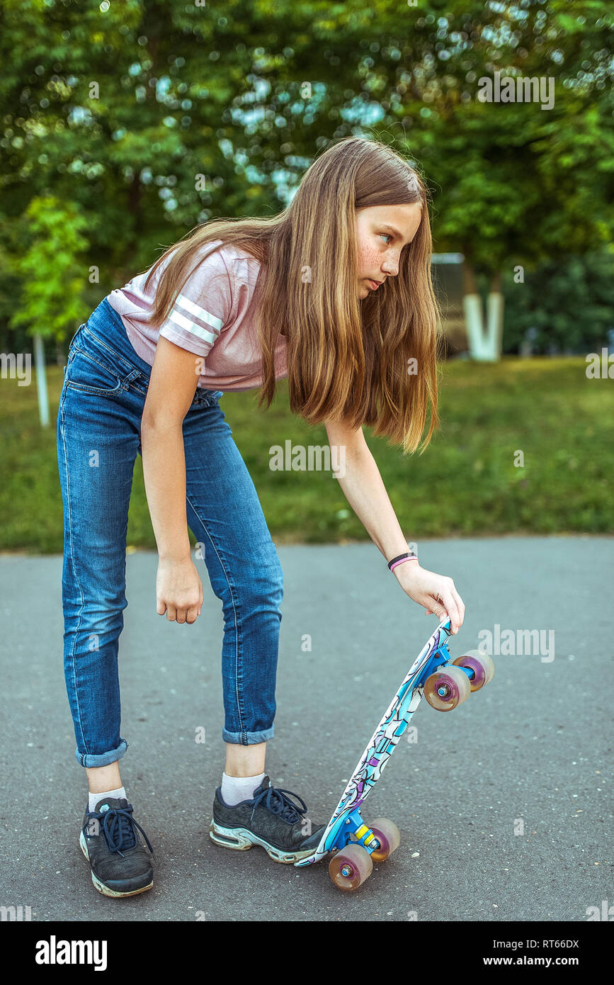 Ragazza adolescente 9-11 anni, equitazione uno skateboard. In estate in città in casual jeans e scarpe da ginnastica. Passeggiata nel parco di estate, dopo scuola Foto Stock