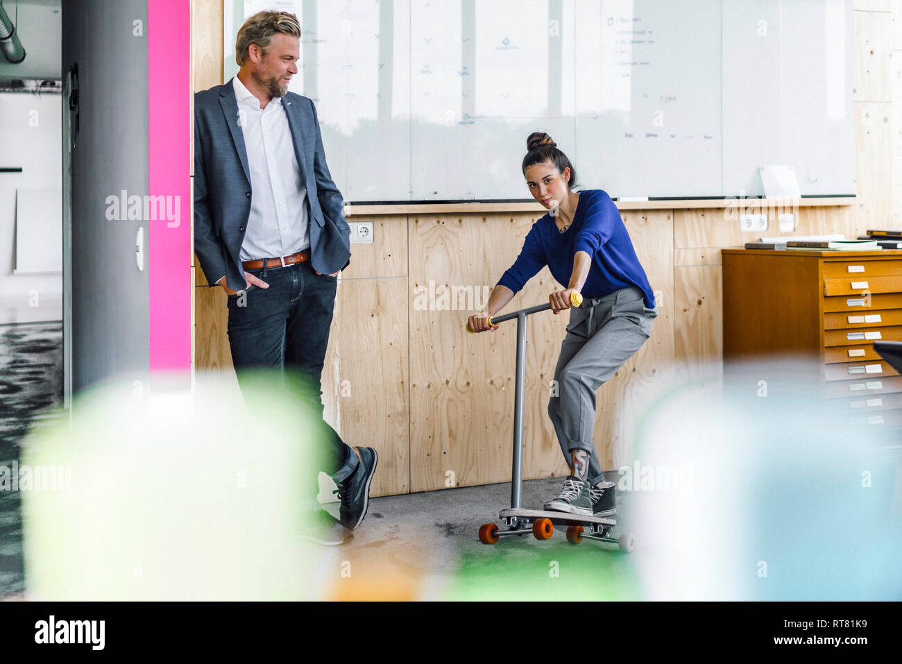 Uomo maturo e il suo assistente a giocare con gli scooter, stando in ufficio nella parte anteriore del pannello bianco Foto Stock