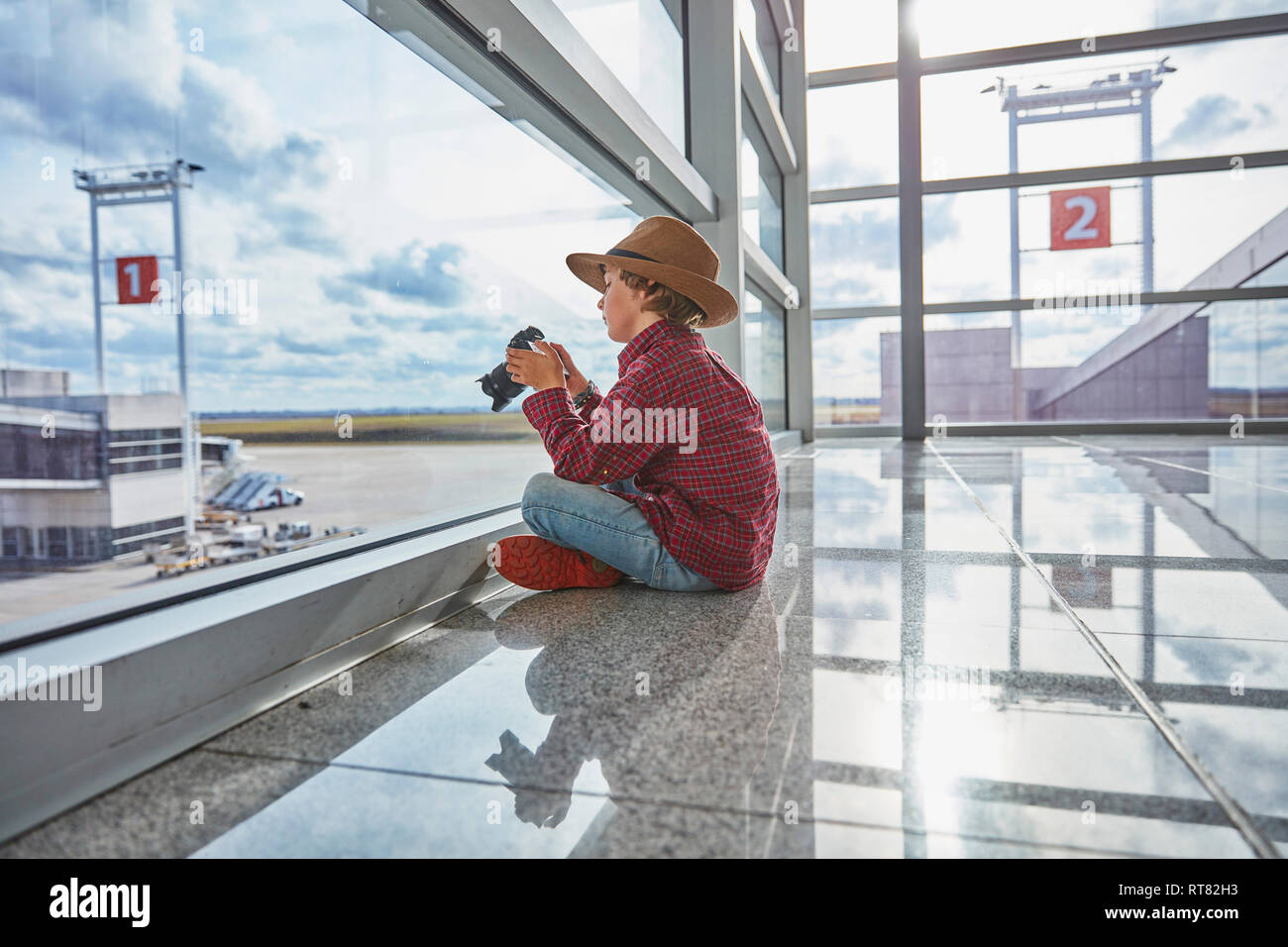 Ragazzo seduto dietro il vetro di finestra all'aeroporto tenendo una fotocamera Foto Stock