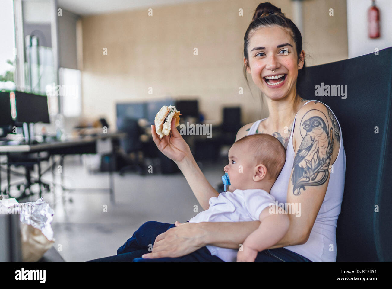 Giovane madre con tatuaggi seduto in ufficio con il suo bambino sul giro, mangiare un panino Foto Stock