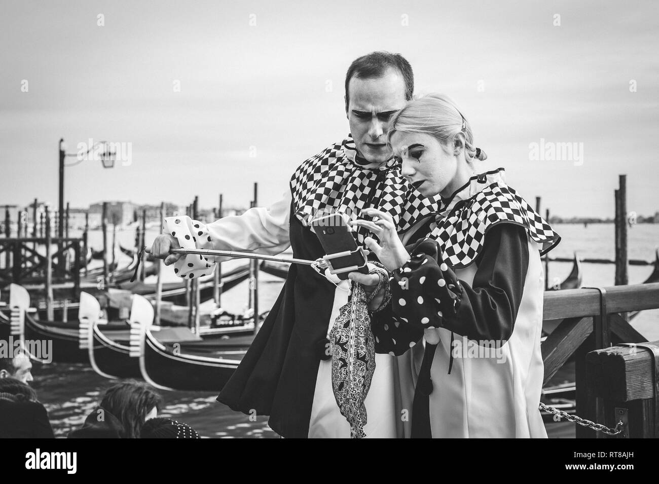 Paio di maschere con un telefono durante il carnevale di Venezia, in piedi vicino alla laguna in un ritratto in bianco e nero Foto Stock