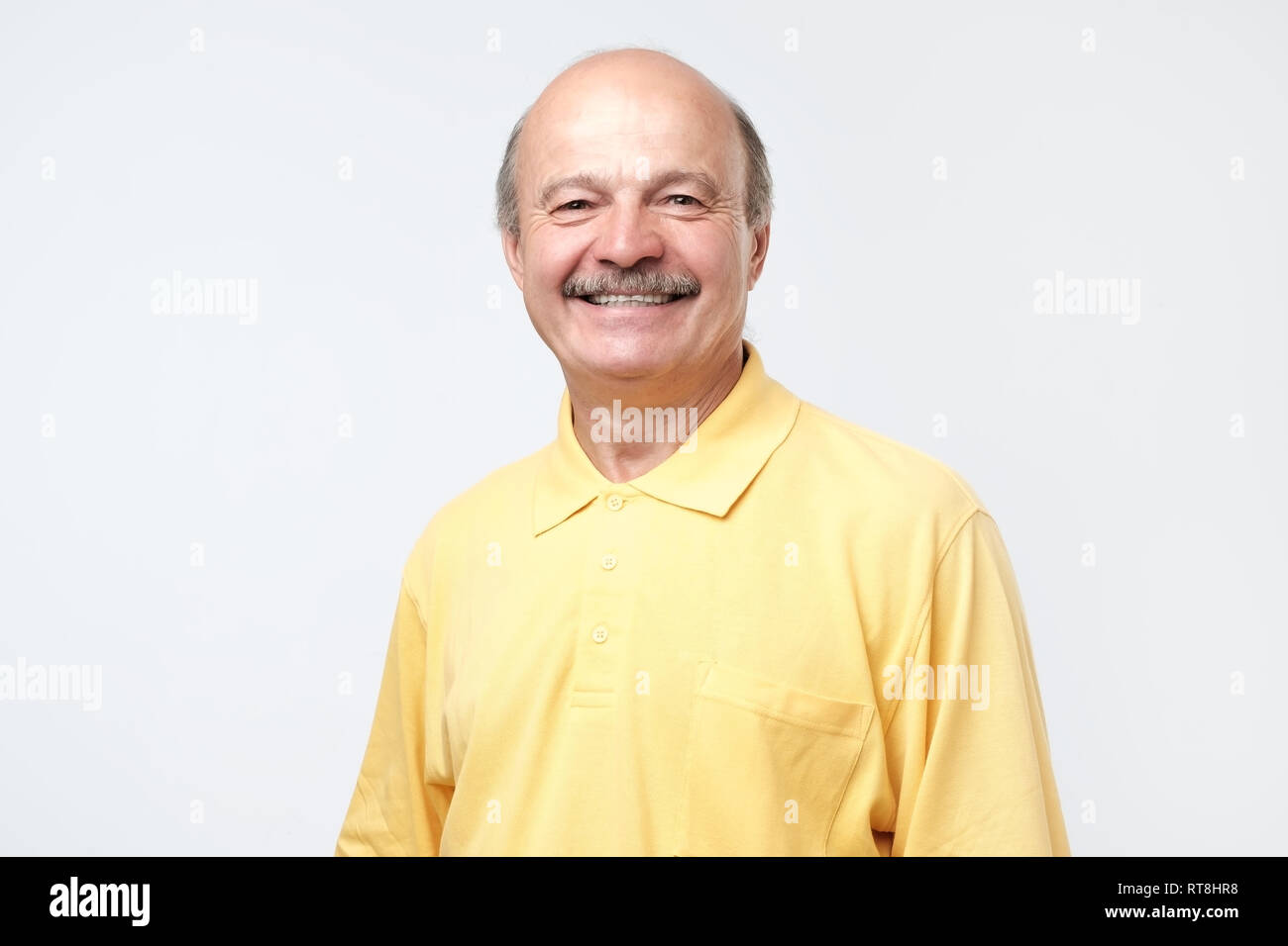 Interessanti spagnolo uomo calvo con i baffi in maglietta gialla grinning al racconto divertente nascondere il dolore. Non sorriso naturale nozione Foto Stock