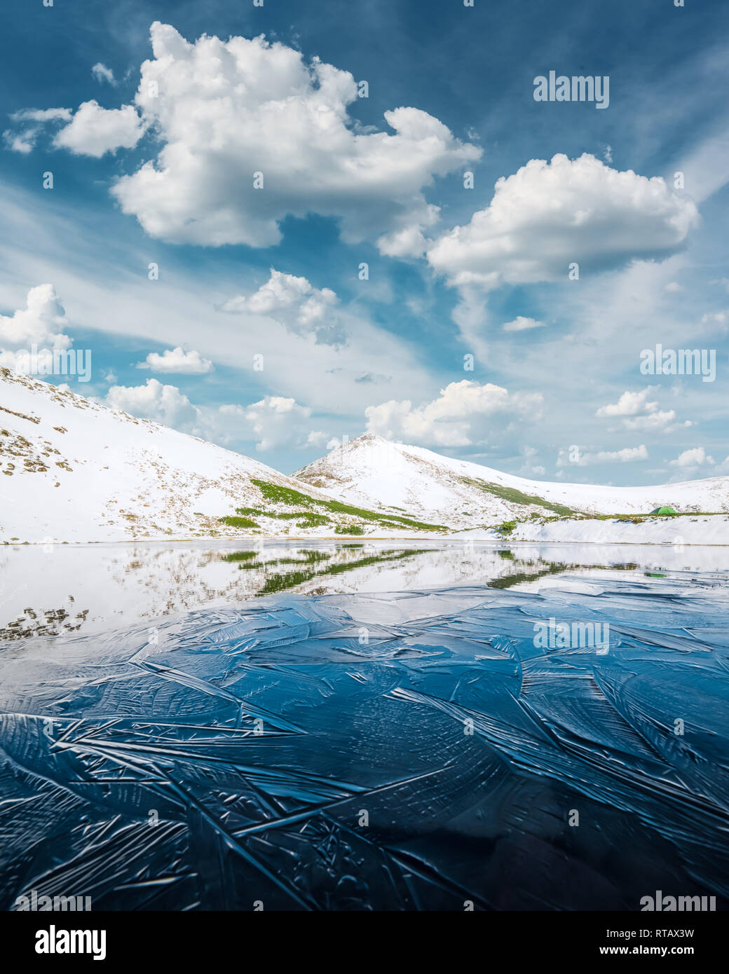 Congelati lago di montagna con il blu ghiaccio e crepe sulla superficie. Il pittoresco paesaggio invernale con colline innevate sotto un azzurro cielo nuvoloso Foto Stock