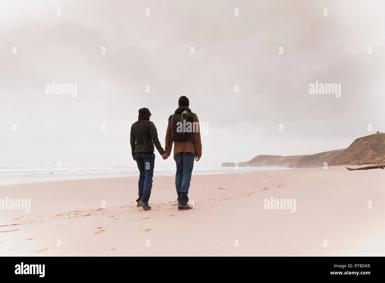 Vista posteriore della coppia giovane con zaino in Warm wear azienda ogni altre mani sulla spiaggia di sabbia vicino al mare e colline Foto Stock