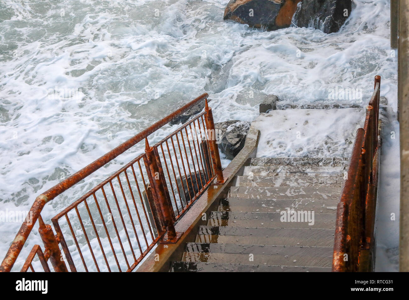 Onde che si infrangono sulle rocce e la preparazione di schiuma fino vecchio arrugginito ringhiera in metallo e passi concreti a Dunedin Beach Nuova Zelanda Isola del Sud Foto Stock